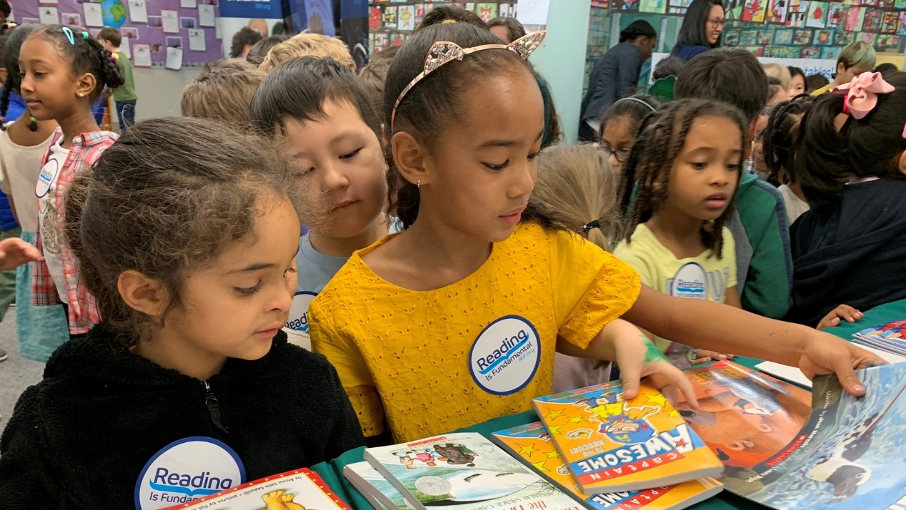 Nestle and RIF Children pick out books at an event organized by Reading Is Fundamental and Nestle. A girl in a yellow shirt with a golden headband stands at the front of the table, reaching out to pick a specific title. Two nearby children look over her shoulder at the spread of books.